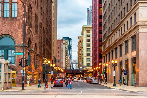 Pedestrians and cars move through a busy downtown Chicago crosswalk lined with tall buildings and streetlights.